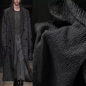 May include: A long, textured, charcoal-colored overcoat with a double-breasted design. The coat is worn over a gray shirt and pants. The fabric has a unique, rippled pattern. A black handbag is held in the hand.