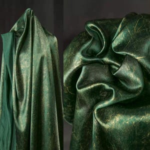 May include: Deep emerald green fabric with a gold metallic pattern. The fabric is artfully draped, creating folds and shadows that highlight its texture and sheen. The overall effect is luxurious.