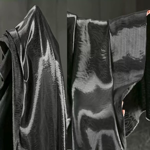 May include: Black fabric with a subtle, repeating pattern. The fabric is draped, creating folds and shadows that highlight its texture and sheen. The material appears to be lightweight and flowing, suitable for apparel or home decor.