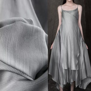 Peut inclure: Une robe grise fluide avec des bretelles fines et une jupe à plusieurs niveaux. Le tissu a une texture subtile et se drape avec élégance. La robe est portée par une personne, avec un gros plan du tissu à gauche.