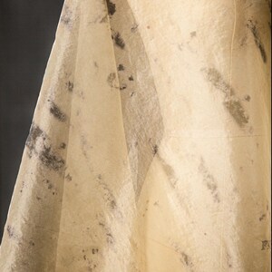 May include: A draped, off-white fabric with dark gray and black mottled patterns. The fabric is draped over a mannequin, creating soft folds and shadows. The material appears to be thin and translucent, suggesting a delicate textile.