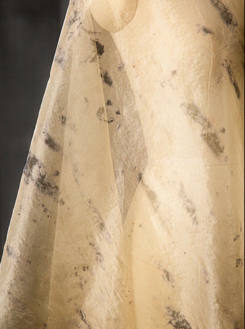 May include: A draped, off-white fabric with dark gray and black mottled patterns. The fabric is draped over a mannequin, creating soft folds and shadows. The material appears to be thin and translucent, suggesting a delicate textile.