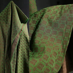 May include: Green fabric with a repeating brown pattern. The fabric is draped, showing the texture and design. The edges are trimmed with a brown border.