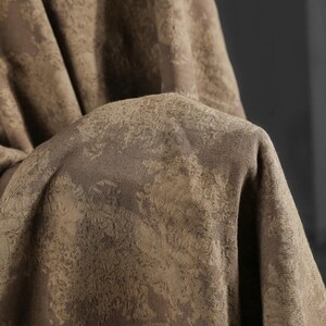May include: Close-up of a folded fabric with a textured, mottled pattern in shades of brown and tan. The fabric appears to be thick and possibly woven, with a subtle sheen. The overall effect is one of depth and visual interest.