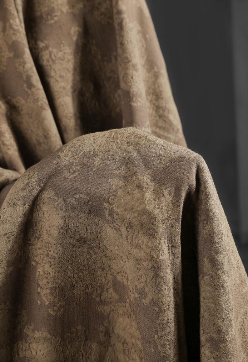 May include: Close-up of a folded fabric with a textured, mottled pattern in shades of brown and tan. The fabric appears to be thick and possibly woven, with a subtle sheen. The overall effect is one of depth and visual interest.