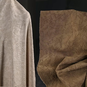 May include: Two pieces of textured fabric in neutral tones. One is a light beige, draped and folded, while the other is a darker brown with a similar embossed pattern. The fabrics appear to be upholstery or drapery material.