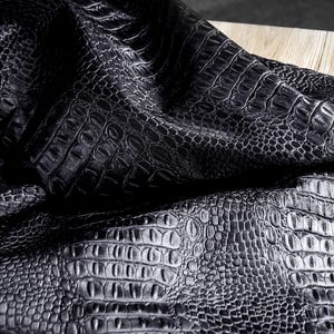 May include: Close-up of black faux crocodile leather fabric. The fabric has a textured surface with a repeating pattern of scales and ridges, creating a realistic crocodile skin appearance. The fabric is draped over a wooden surface.