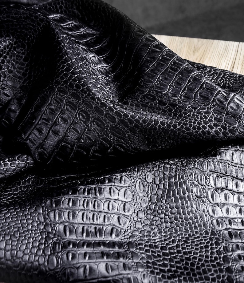 May include: Close-up of black faux crocodile leather fabric. The fabric has a textured surface with a repeating pattern of scales and ridges, creating a realistic crocodile skin appearance. The fabric is draped over a wooden surface.