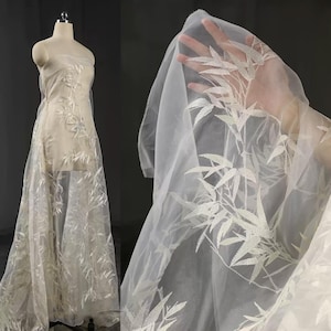 May include: Delicate white fabric featuring an embroidered bamboo leaf pattern. The fabric is draped over a dress form and held up to show the transparency and detail of the leaf design. Suitable for bridal wear or decorative projects.