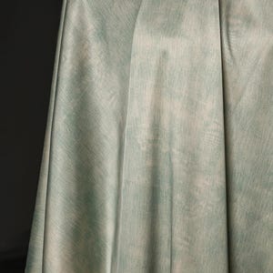May include: A draped fabric in shades of pale green and off-white, with a textured, watercolor-like appearance. The fabric is folded, creating soft shadows and highlighting the subtle color variations. The overall effect is soft and ethereal.
