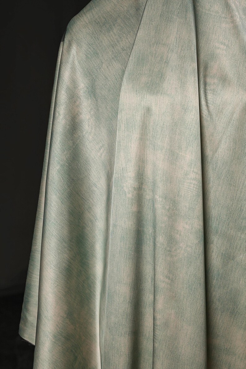 May include: A draped fabric in shades of pale green and off-white, with a textured, watercolor-like appearance. The fabric is folded, creating soft shadows and highlighting the subtle color variations. The overall effect is soft and ethereal.