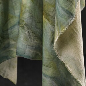 Puede incluir: Primer plano de tela drapeada en tonos verdes y azules, con efecto acuarela. La tela tiene un aspecto texturizado con pinceladas visibles y un borde deshilachado, lo que sugiere una bufanda o textil.