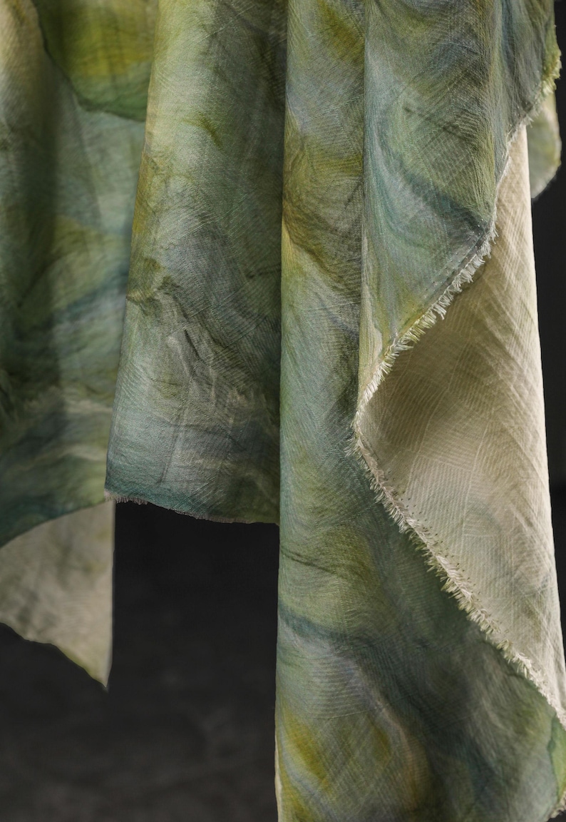 Puede incluir: Primer plano de tela drapeada en tonos verdes y azules, con efecto acuarela. La tela tiene un aspecto texturizado con pinceladas visibles y un borde deshilachado, lo que sugiere una bufanda o textil.