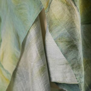 Puede incluir: Primer plano de tela fluida en tonos verdes, amarillos y azules. El material parece ligero, con un patr&oacute;n sutil visible en los pliegues. La tela est&aacute; en capas, creando profundidad y textura, lo que sugiere una bufanda o chal.