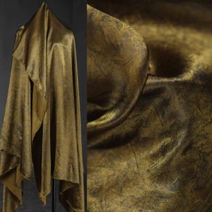 Könnte beinhalten: Ein drapierter Stoff mit metallisch goldener Oberfläche. Das Material hat ein strukturiertes Aussehen mit dunkleren, schattigen Bereichen, die Tiefe erzeugen. Der Stoff ist gefaltet und hängt in weichen, fließenden Falten, was auf ein luxuriöses Textil hindeutet.