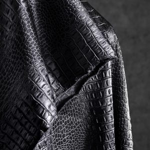 May include: Close-up of black leather fabric with a crocodile skin texture. The material is folded, revealing the intricate pattern and the texture of the material. The lighting highlights the details of the embossed design.