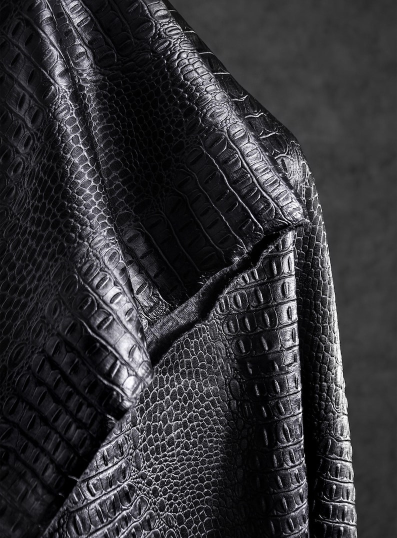 May include: Close-up of black leather fabric with a crocodile skin texture. The material is folded, revealing the intricate pattern and the texture of the material. The lighting highlights the details of the embossed design.