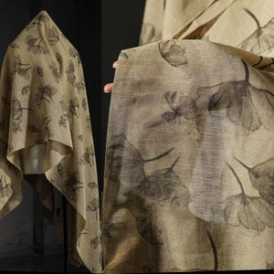 May include: Beige fabric with a floral design in grey tones. The material is draped over a form and held up by a hand, highlighting the pattern and texture. The floral print features large, stylized blooms.