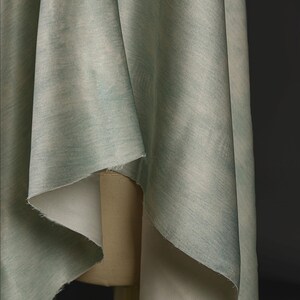 May include: Close-up of draped fabric in shades of pale green and blue. The fabric has a subtle, textured pattern and is folded over a mannequin. The fabric's edges are visible, revealing a lighter, off-white underside. The background is a dark gray.