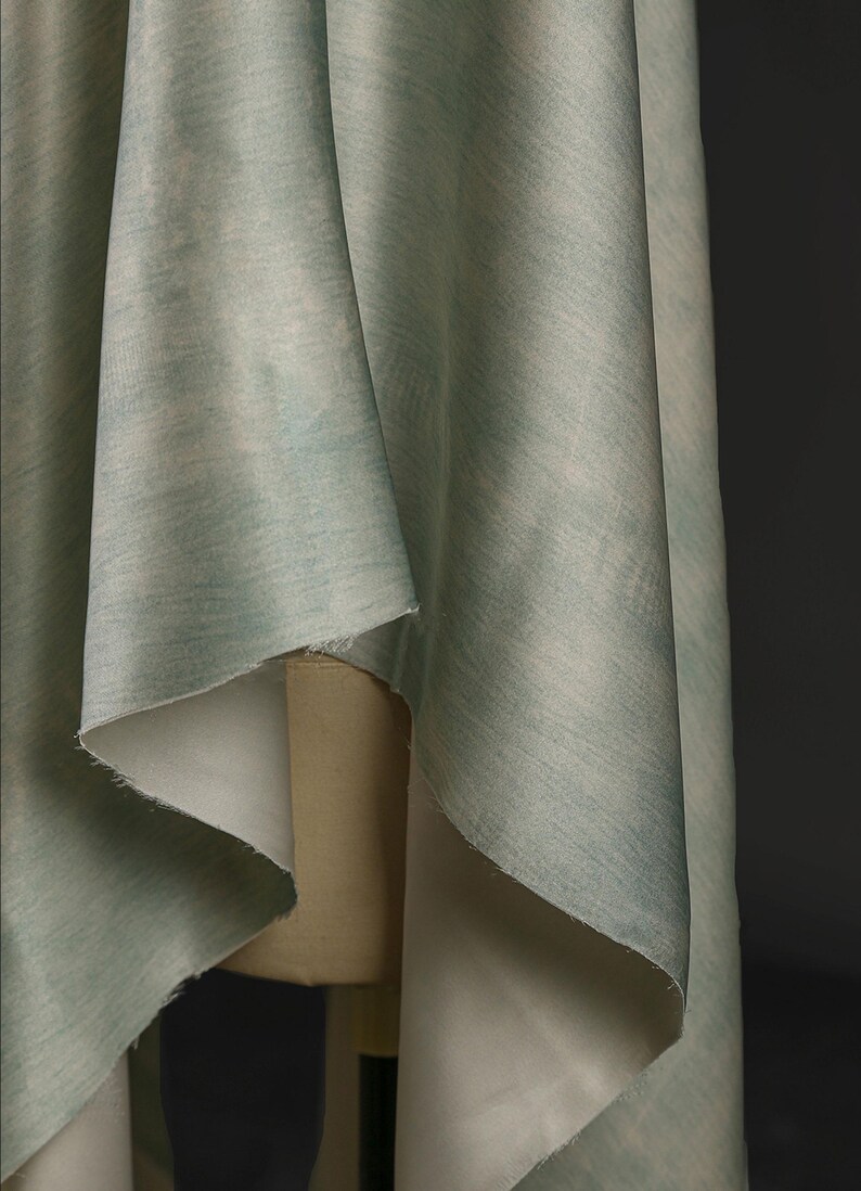 May include: Close-up of draped fabric in shades of pale green and blue. The fabric has a subtle, textured pattern and is folded over a mannequin. The fabric's edges are visible, revealing a lighter, off-white underside. The background is a dark gray.