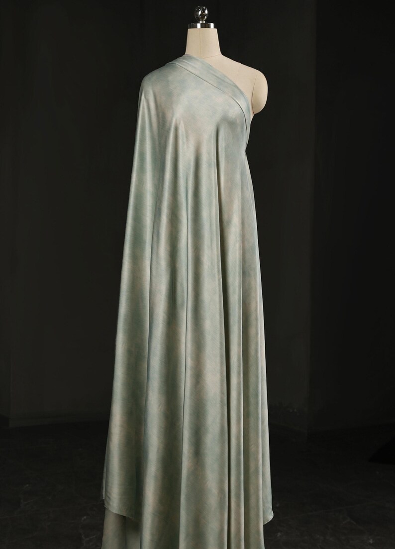 May include: A draped fabric in shades of pale blue and green is displayed on a mannequin. The fabric has a soft, watercolor-like appearance, with subtle variations in color. The material is draped to create a flowing effect, suggesting it is suitable for apparel or home decor.