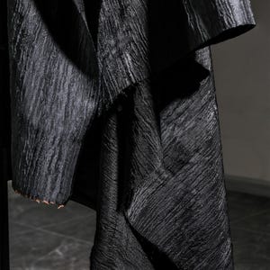 May include: Close-up of a black fabric with a textured, rippled surface. The fabric appears to be draped, with folds and shadows creating depth. The edges of the fabric are visible, showing a raw, unfinished edge.