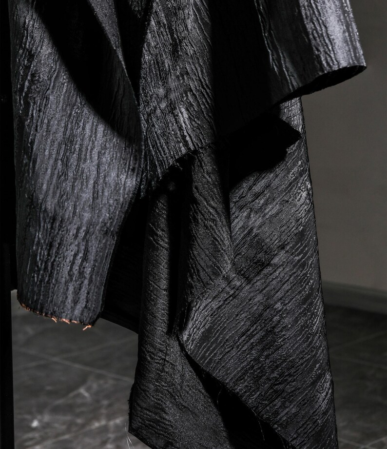 May include: Close-up of a black fabric with a textured, rippled surface. The fabric appears to be draped, with folds and shadows creating depth. The edges of the fabric are visible, showing a raw, unfinished edge.