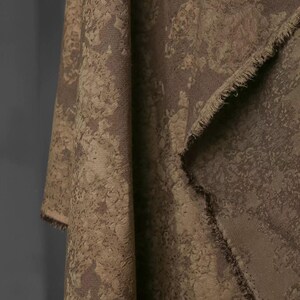 May include: Close-up of a draped fabric with a textured, mottled pattern in shades of brown and tan. The fabric has a raw, unfinished edge, revealing the weave. The overall effect is rustic and organic.