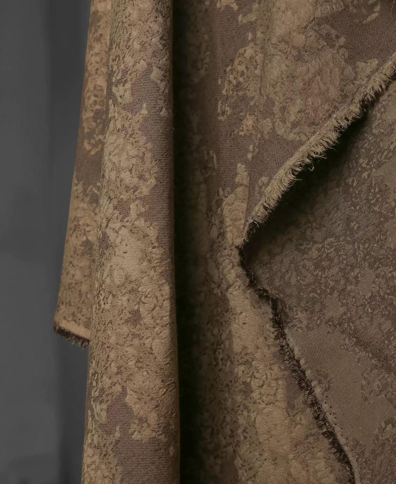 May include: Close-up of a draped fabric with a textured, mottled pattern in shades of brown and tan. The fabric has a raw, unfinished edge, revealing the weave. The overall effect is rustic and organic.