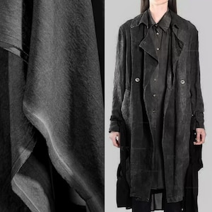 Puede incluir: Un abrigo tipo trench de color gris oscuro con diseño de doble botonadura y solapas grandes. El abrigo se lleva sobre una camisa y un pantalón a juego. La tela tiene un aspecto texturizado. El estilo general es minimalista y moderno.