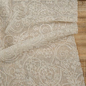May include: Close-up of ivory lace fabric with a detailed floral pattern. The delicate material is draped over a wooden surface, showcasing the intricate design and texture. The lace is a light cream color.