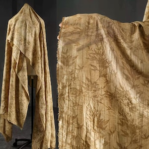 May include: Tan fabric with a repeating leaf pattern. The fabric is draped over a mannequin and also held up to show the texture and detail. The fabric has a crinkled appearance and a raw edge.