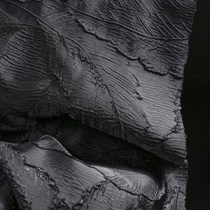 Op de afbeelding: Close-up van een zwarte stof met een bladachtig patroon. De stof heeft een gelaagde uitstraling met gedetailleerde nerven en een licht verhoogde textuur, wat een gevoel van diepte en dimensie cre&euml;ert. De algehele esthetiek is donker en elegant.
