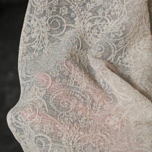 May include: Close-up of a cream-colored, intricately patterned fabric. The design features swirling floral and paisley motifs, creating a delicate, textured appearance. The fabric appears to be lightweight and semi-transparent, with a soft, flowing drape.