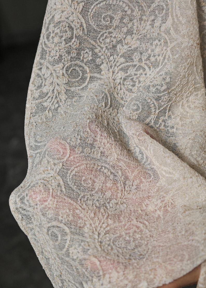 May include: Close-up of a cream-colored, intricately patterned fabric. The design features swirling floral and paisley motifs, creating a delicate, textured appearance. The fabric appears to be lightweight and semi-transparent, with a soft, flowing drape.