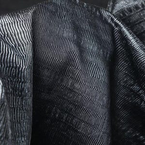 May include: Close-up of a dark fabric with a textured, ribbed pattern. The fabric appears to be folded, revealing the texture and sheen of the material. The color is a deep, dark shade, possibly black or navy.