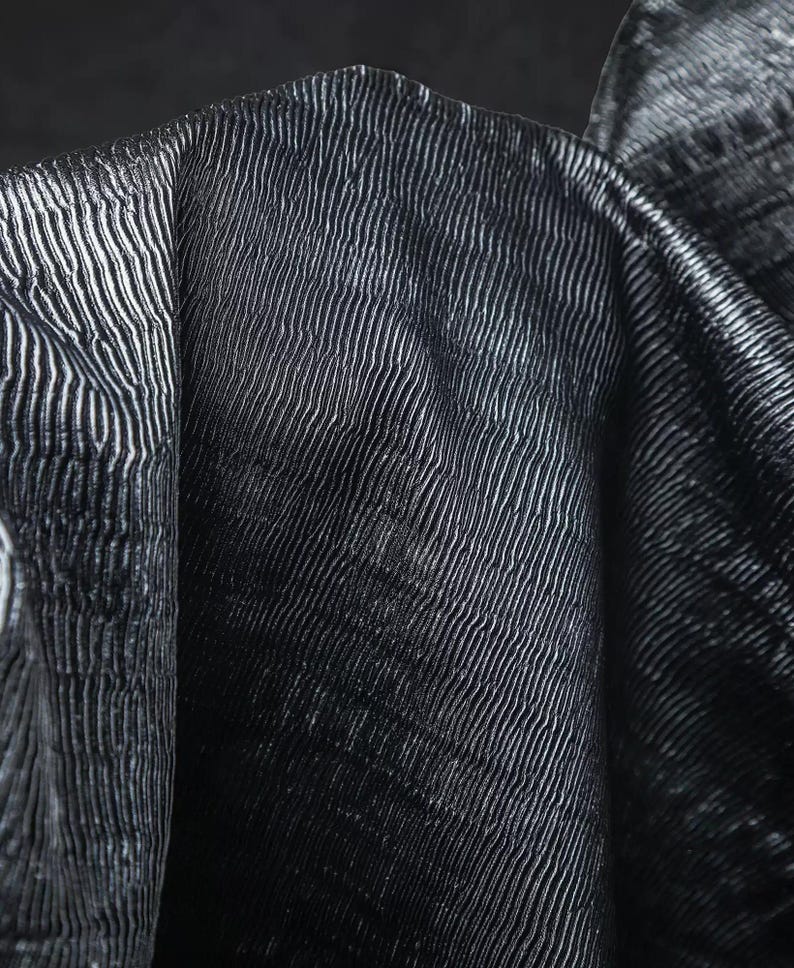 May include: Close-up of a dark fabric with a textured, ribbed pattern. The fabric appears to be folded, revealing the texture and sheen of the material. The color is a deep, dark shade, possibly black or navy.