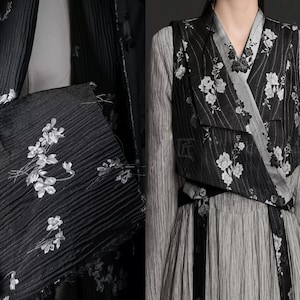 Puede incluir: Tela negra plisada con bordado floral blanco. La tela forma parte de una prenda. Un chaleco negro con estampado floral blanco está superpuesto a un vestido gris. La estética general es elegante y detallada.