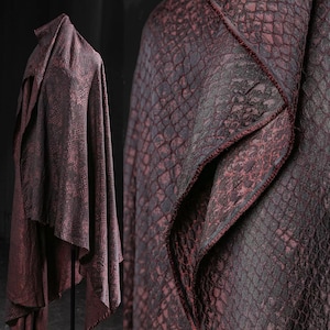 Pode incluir: Tecido drapeado com padrão de pele de cobra em tons de bordô e preto. O tecido é exibido em um manequim e também em uma vista em close-up, mostrando a textura e o design. As bordas são finalizadas com uma borda vermelha contrastante.