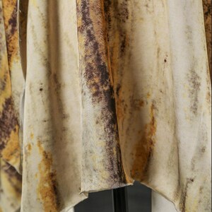 May include: Close-up of a flowing fabric with a mottled pattern. The fabric features a blend of beige, brown, and yellow hues, creating a textured, organic appearance. The material drapes in soft folds, suggesting a lightweight textile.