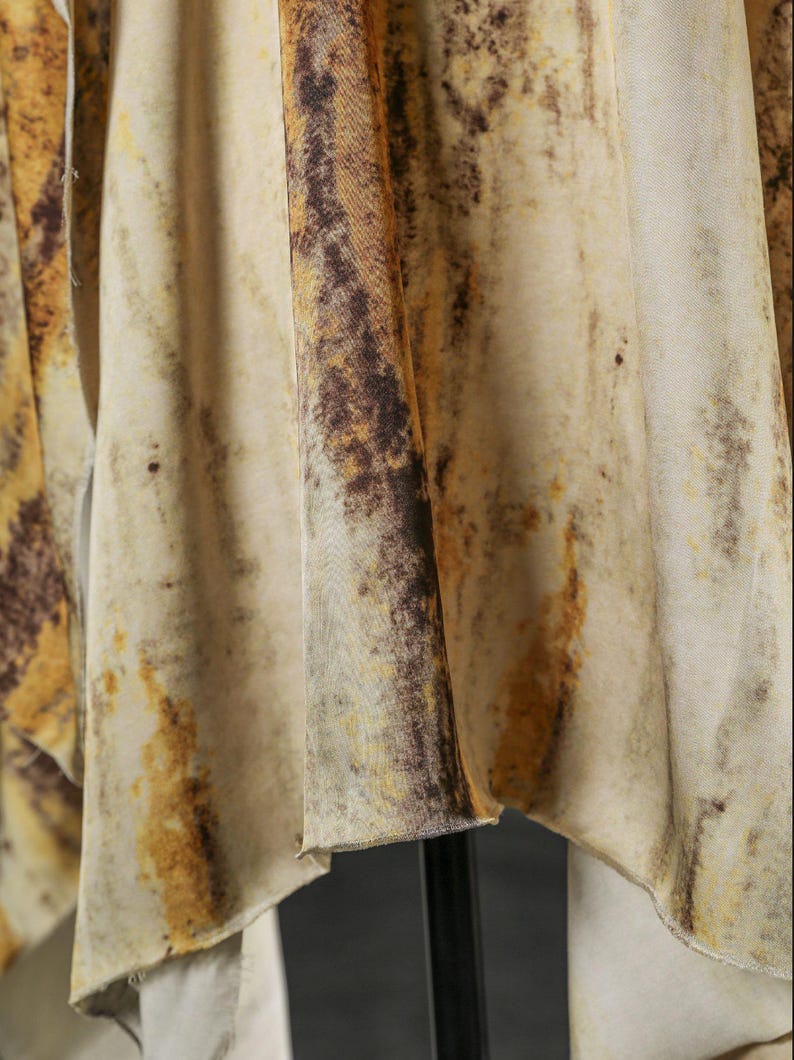 May include: Close-up of a flowing fabric with a mottled pattern. The fabric features a blend of beige, brown, and yellow hues, creating a textured, organic appearance. The material drapes in soft folds, suggesting a lightweight textile.