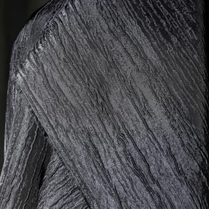 May include: Close-up of a textured black fabric with a crinkled appearance. The fabric has a subtle sheen and a pattern of vertical lines, creating a sense of depth and dimension. The folds and angles of the fabric are visible.