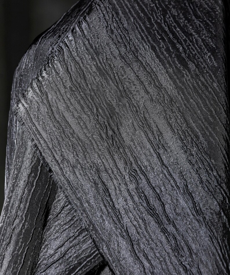 May include: Close-up of a textured black fabric with a crinkled appearance. The fabric has a subtle sheen and a pattern of vertical lines, creating a sense of depth and dimension. The folds and angles of the fabric are visible.