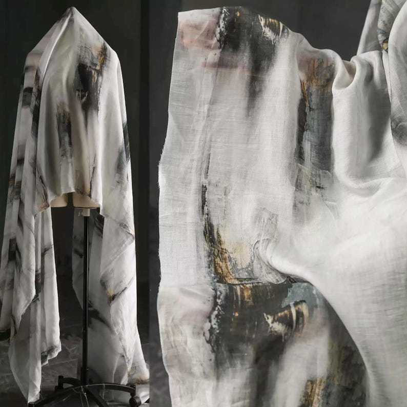 May include: White fabric featuring an abstract design in black, grey, and brown tones. The material is draped over a dress form and also shown in a close-up, showcasing the texture and pattern.