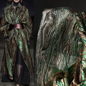 Könnte beinhalten: Ein langer Mantel mit einem metallischen, wirbelnden Muster in Grün-, Braun- und Goldtönen. Der Mantel ist mit einem breiten, dunkelbraunen Gürtel in der Taille zusammengebunden. Der Stoff hat ein strukturiertes, geripptes Aussehen.