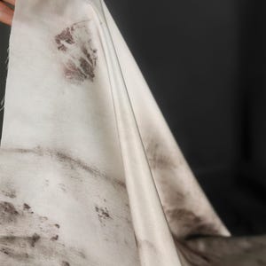 May include: Off-white fabric with dark brown and maroon botanical prints. The fabric is draped, revealing folds and textures. The dark background contrasts with the light fabric, highlighting the organic patterns.