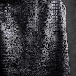 May include: Close-up of a black leather jacket with a crocodile skin texture. The jacket's surface is detailed with a repeating pattern of scales, creating a textured and luxurious appearance. The dark color and pattern suggest a bold and stylish design.