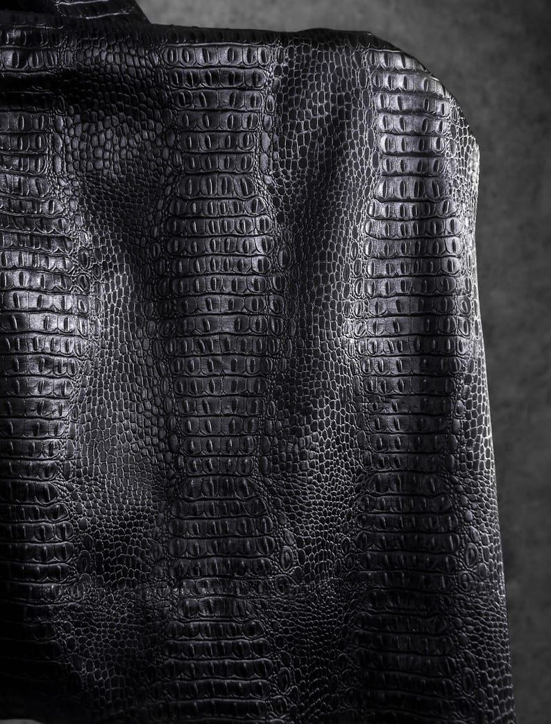 May include: Close-up of a black leather jacket with a crocodile skin texture. The jacket's surface is detailed with a repeating pattern of scales, creating a textured and luxurious appearance. The dark color and pattern suggest a bold and stylish design.