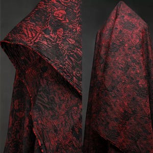 Puede incluir: Una capa con capucha hecha de tela con un patrón floral rojo y negro. La tela está drapeada, mostrando la textura y el diseño. La capucha es visible, mostrando los intrincados detalles del patrón. La tela es texturizada.