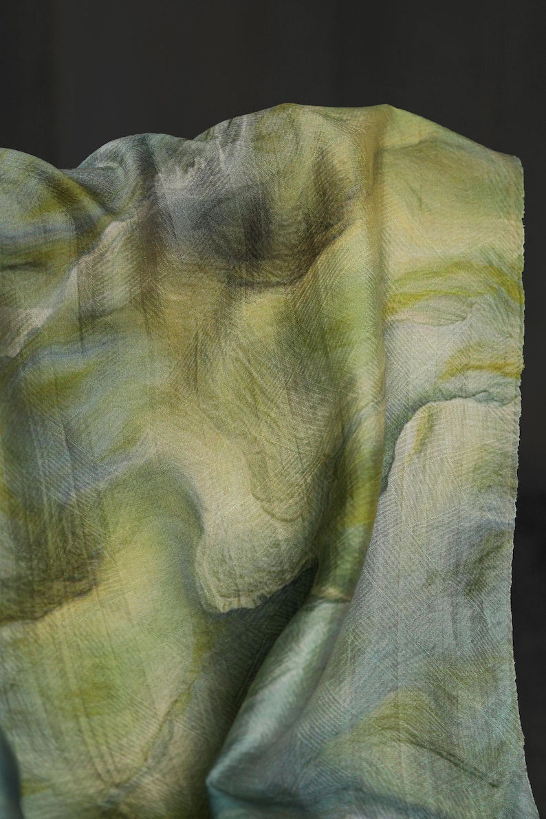 Puede incluir: Una tela drapeada con un patr&oacute;n similar a una acuarela en tonos verdes, amarillos y azules. El textil tiene una apariencia texturizada, con detalles tejidos visibles. Los pliegues crean una sensaci&oacute;n de profundidad y movimiento.